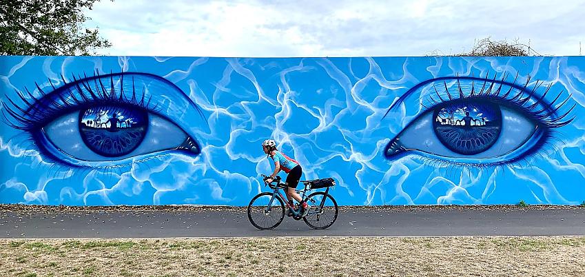 A woman is cycling past a big wall mural of two eyes; there's a figure reflected in the two eyes, it's painted in different shades of blue. The woman is on a touring bike that's packed ready for touring. She is wearing cycling shorts and jersey