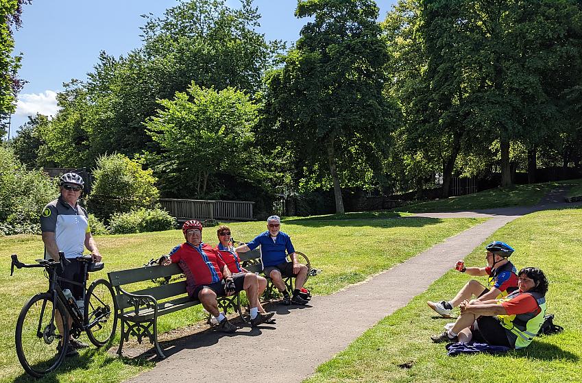 A group of cyclists is taking a break in a park. They are sitting on benches and on the ground, enjoying a drink and a rest