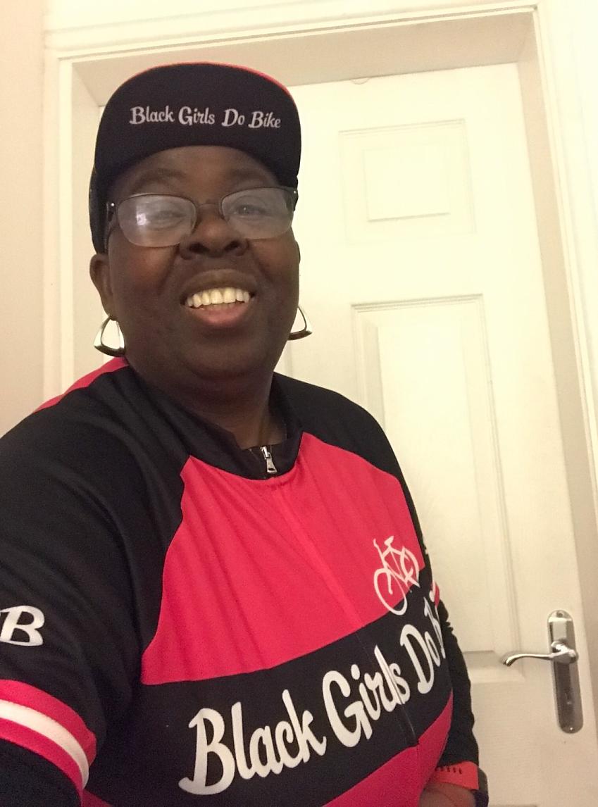 A woman is smiling at the camera. She is wearing a red and black baseball cap with the slogan Black girls do bike. She's also wearing a matching cycling jersey.
