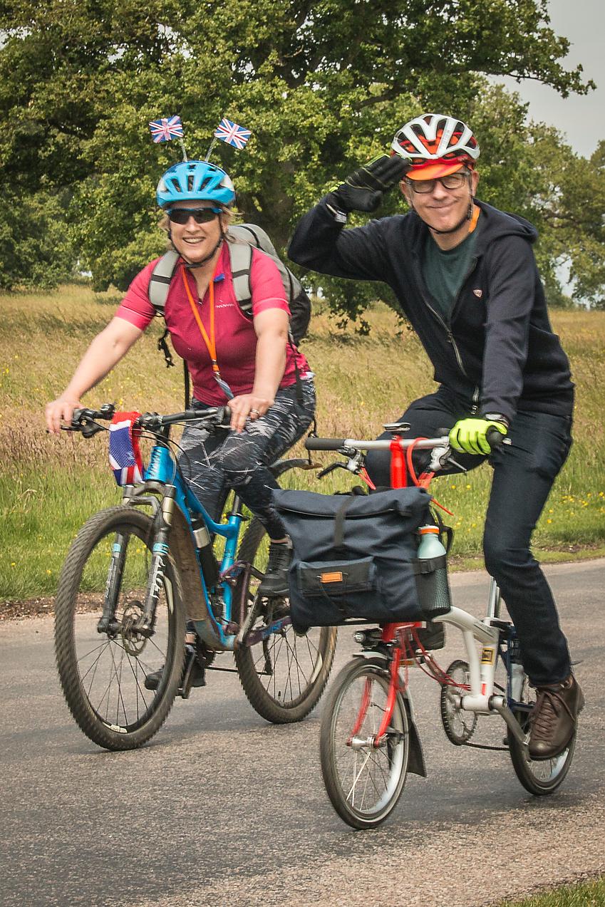 Two people are cycling along a road. A man is on a Brompton and is saluting. A woman is on a mountain bike and she has two small Union Jack flats sticking out of her helmet, there's also a red, white and blue headband on her handlebar