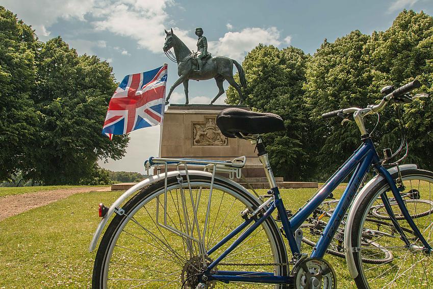 A blue hybrid bike with a Union Jack flag attached to the rear rack is propped up on grass in front of a statue of Queen Elizabeth on a horse. There's also a black bike lying on the grass