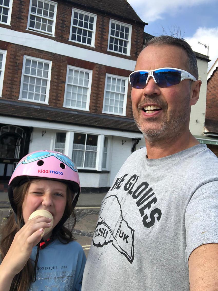 A young girl wearing a pink cycle helmet eating an ice cream with her father, stood outside a historic building