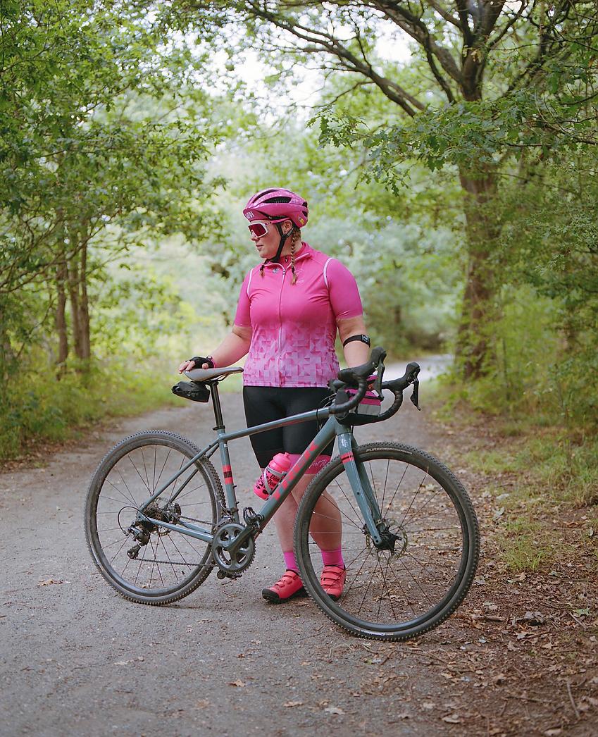 The same woman, wearing the same outfit but this time with a matching pink helmet and sunglasses, stands with a grey and pink gravel bike