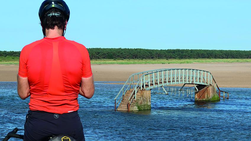 A man stands in the foreground, his back to the camera. He's wearing a bright orange jersey and a black helmet. In the background is a bridge in the middle of a river