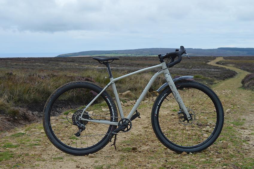 Mason InSearchOf Force 1X, a chunky, grey  gravel bike that looks like a cross between a mountain bike and road bike, with drop bars and fat tyres, propped up on a gravel and grass path