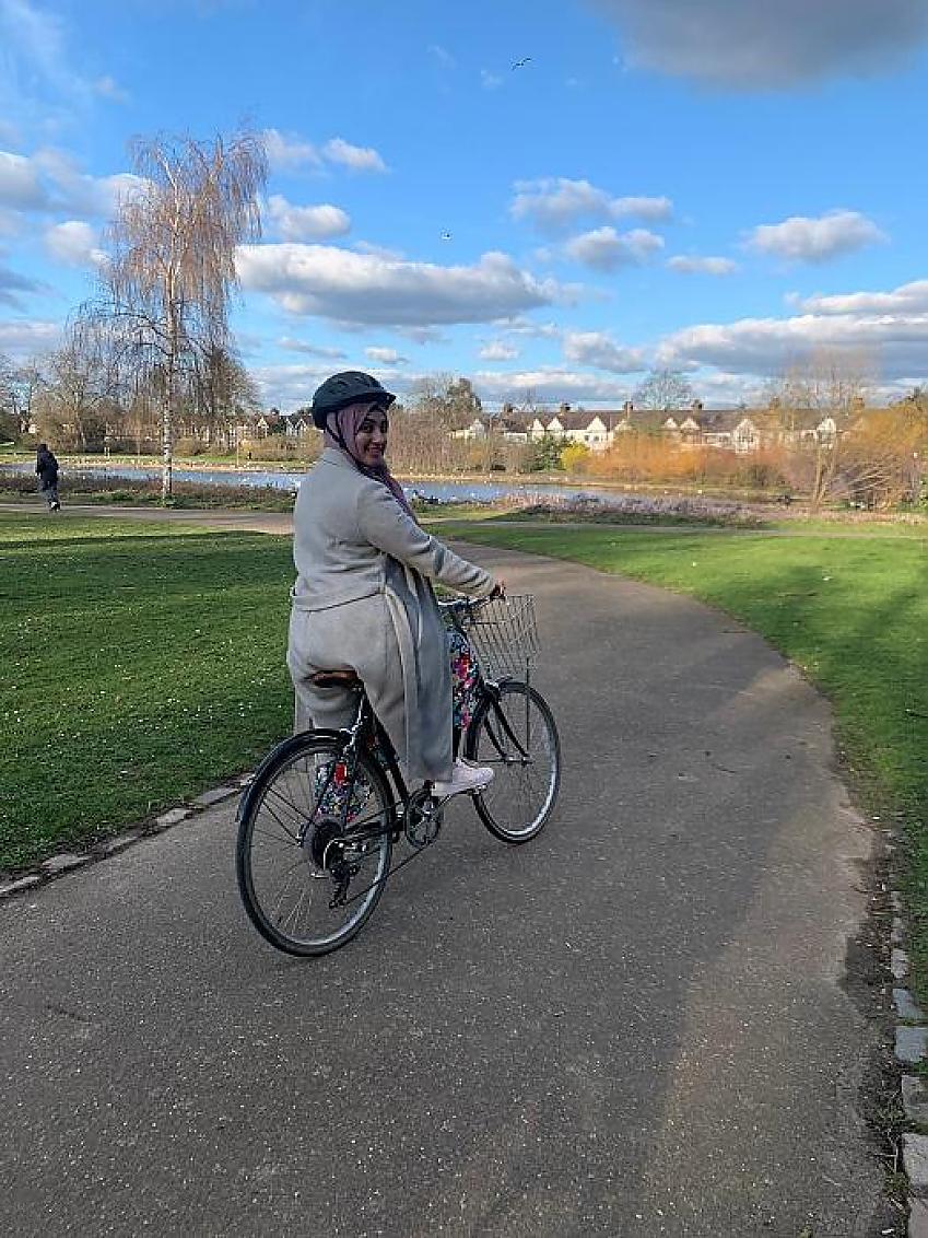 A woman smiles at us as she poses on her bicycle along a tarmac path in a country park. She is wearing a helmet and a long grey coat.