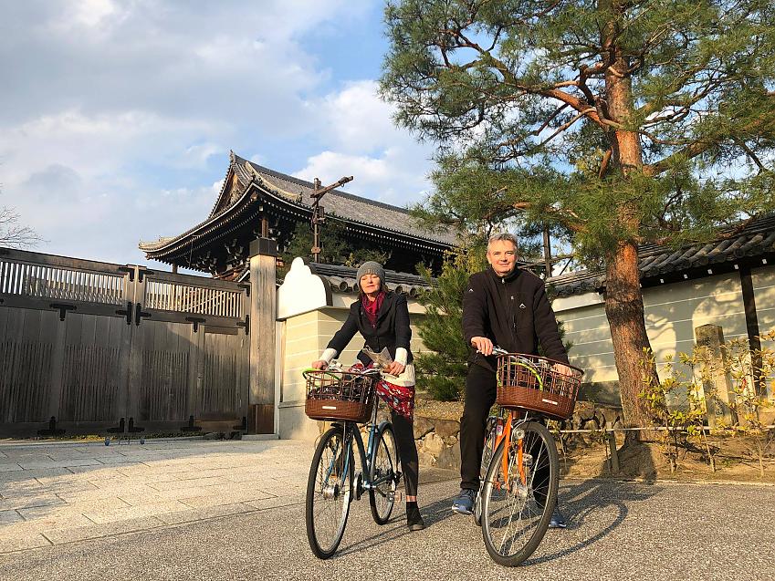 A man and woman are seated on their hire bikes; his is orange, hers is blue. They both have baskets on the front. Both people are wearing normal clothes. There's a Shinto temple in the background