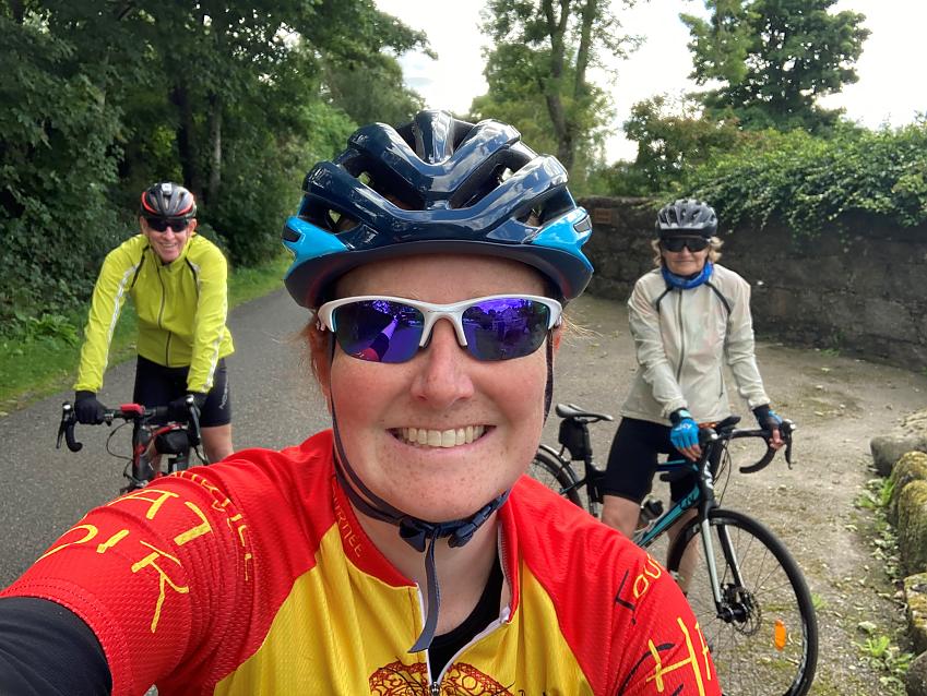 A woman, Naomi, wearing cycling clothing and broadly smiling, is taking a selfie, with her parents parents in the background on road bikes, in a rural setting
