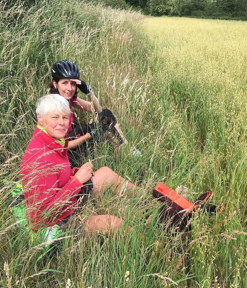 Two women are in a field having lunch. They're dressed in cycling clothing and smiling at the camera