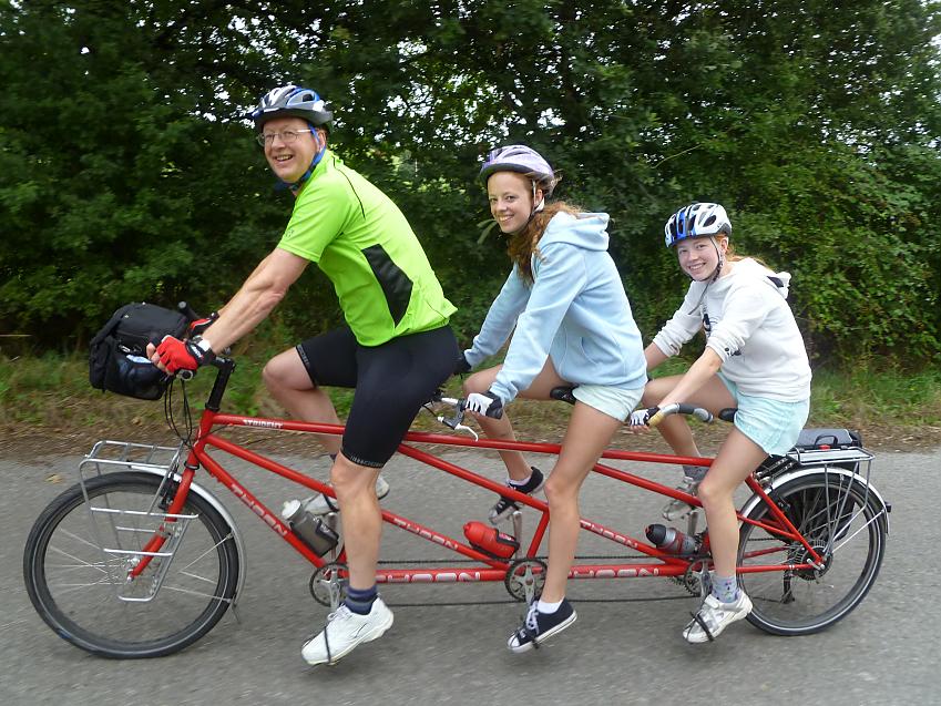 Three cyclists are riding a red tandem for three people. A man is in front followed by two younger girls
