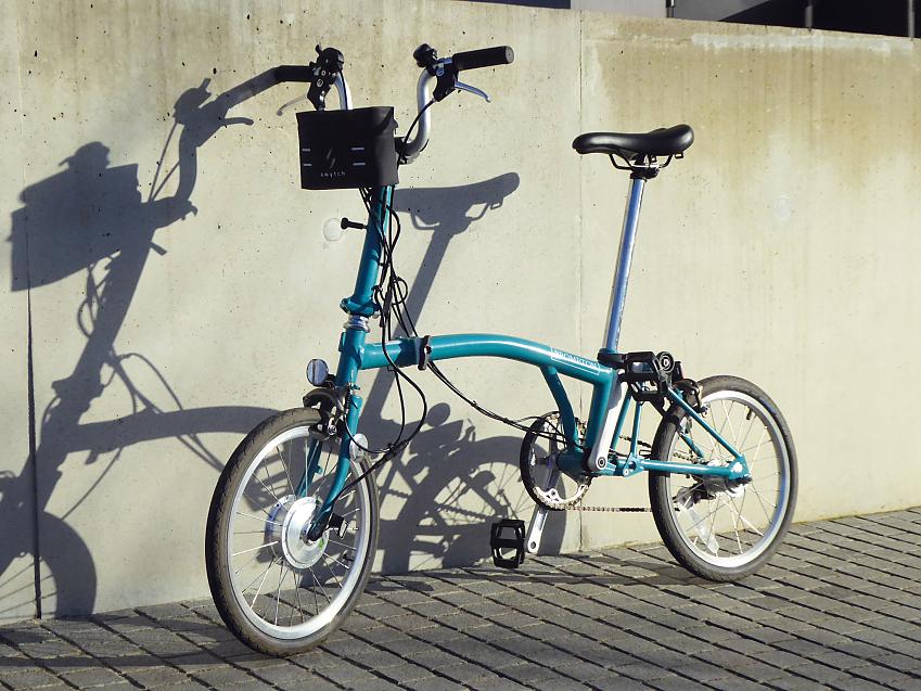 A light blue Brompton bike leaning against a wall. It has an electric battery pack attached to the handlebar and the front wheel has a hub motor