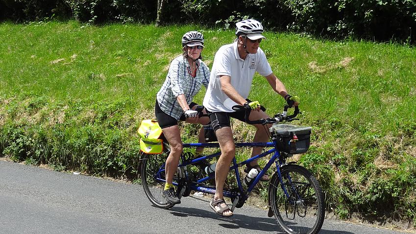 Two cyclists are riding a blue tandem. This time a women takes the back seat, with a man in front