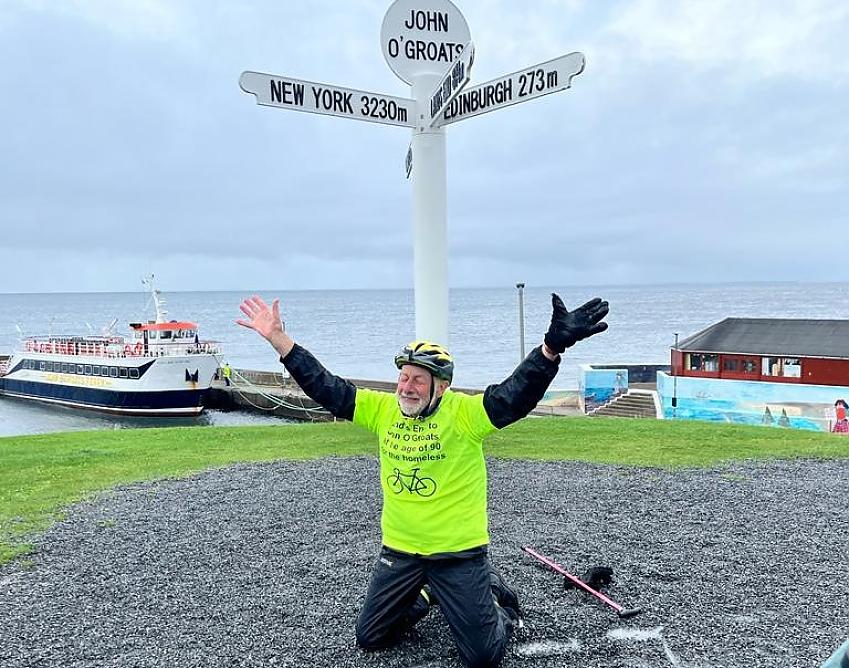 A man kneeling on the ground under the signpost at John O'Groats