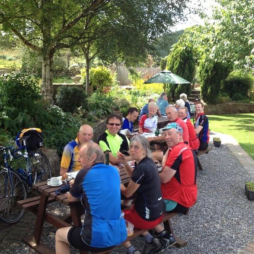 A group of cyclists are sitting at outside tables as they enjoy a cup of tea and some cake