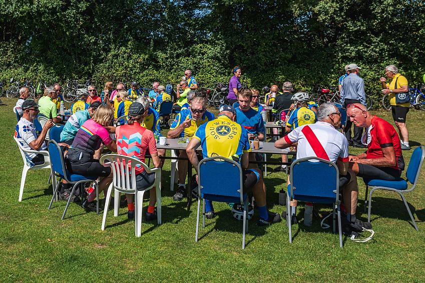 A group of people all in cycling gear are sitting at big tables outside on the grass. They are eating and drinking. Lots of bikes can be seen propped against a hedge in the background