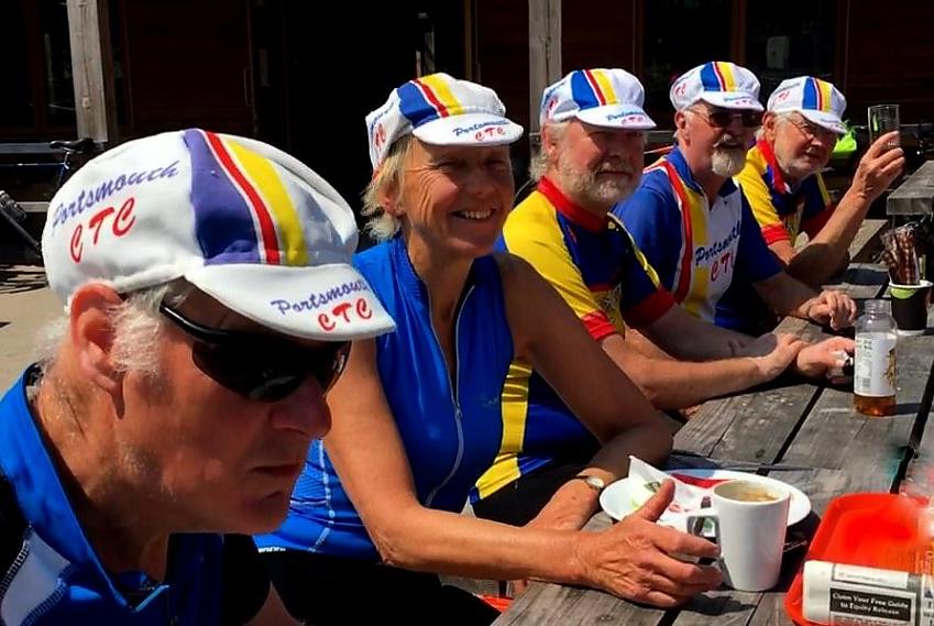 A group of older cyclists are seated at an outdoor table. They are enjoying drinks and a bite to eat. They are all wearing cycling jerseys and caps in the blue and gold colours of Portsmouth CTC
