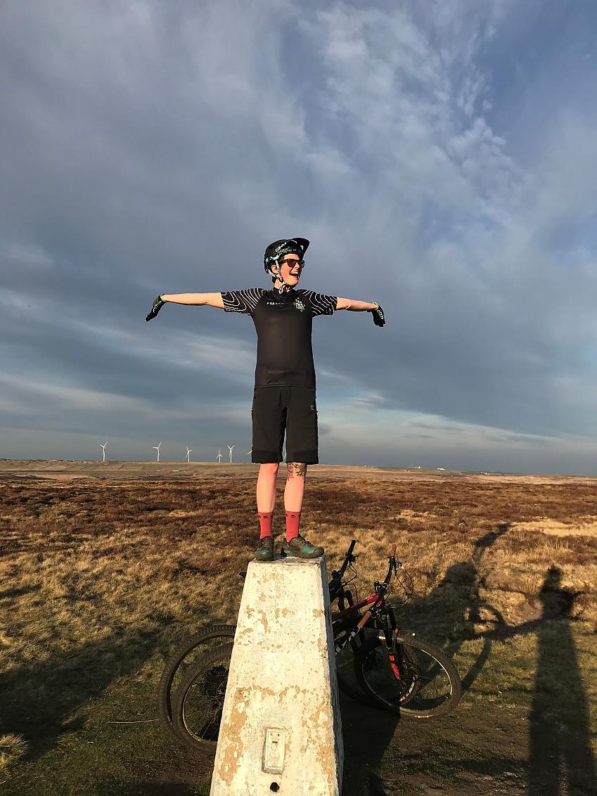 A woman is standing on a concrete plinth set in a large, grassy field. In the background are wind turbines. She is has her arms wide and looks very pleased. She's wearing black cycling clothing; a couple of bikes are leaning against the plinth