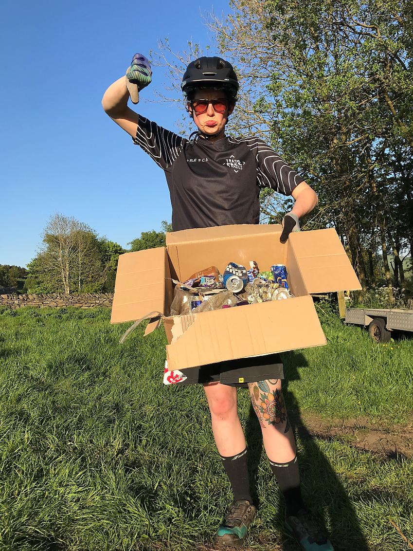 A woman pulling an unhappy face and showing a thumbs down is holding a cardboard box full of rubbish. She is wearing a black Trash Free Trails cycling jersey, a black cycling helmet and sunglasses