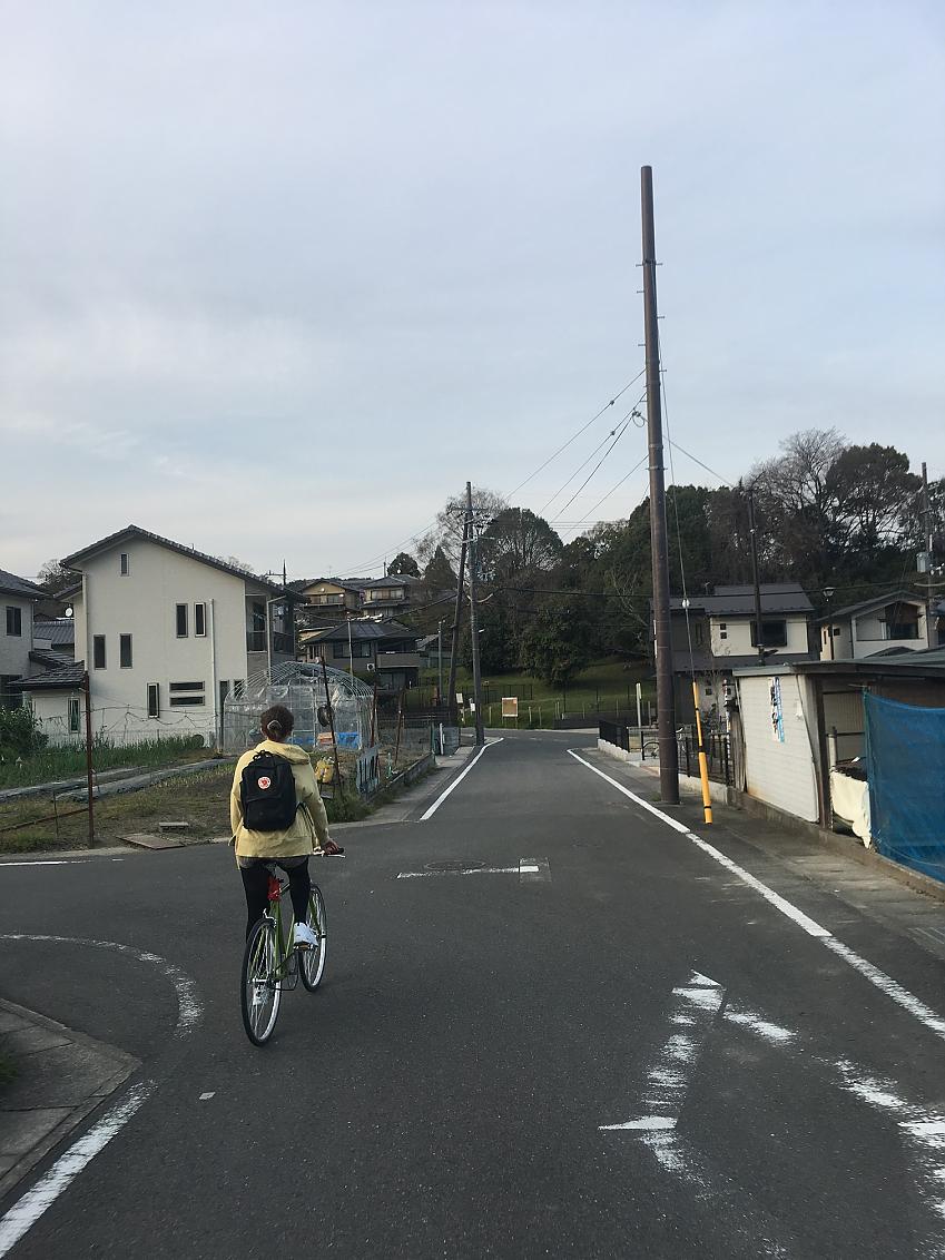 A woman is cycling on a green bike on a quiet road in Kyoto, Japan. She is heading away from the camera; she is wearing a yellow jacket and has a rucksack on her back