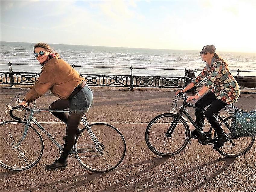 Two women are riding along the seafront. One is wearing a suede jacket and shorts and riding a silver road bike, the other is wearing a floral shirt and jeans and riding a black hybrid bike