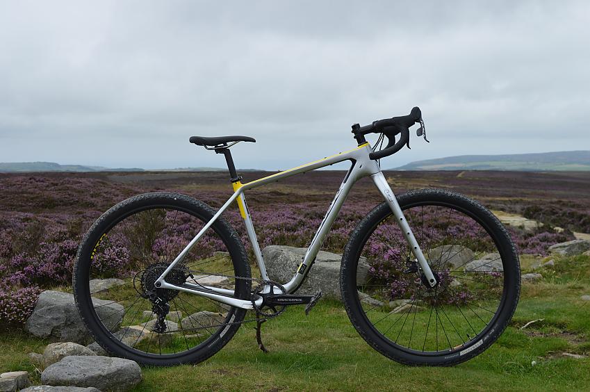 Salsa CutThroat C Apex 1, a grey and yellow gravel bike with drop bars and fat tyres, propped up at the top of a hill with lots of purple heather in the background