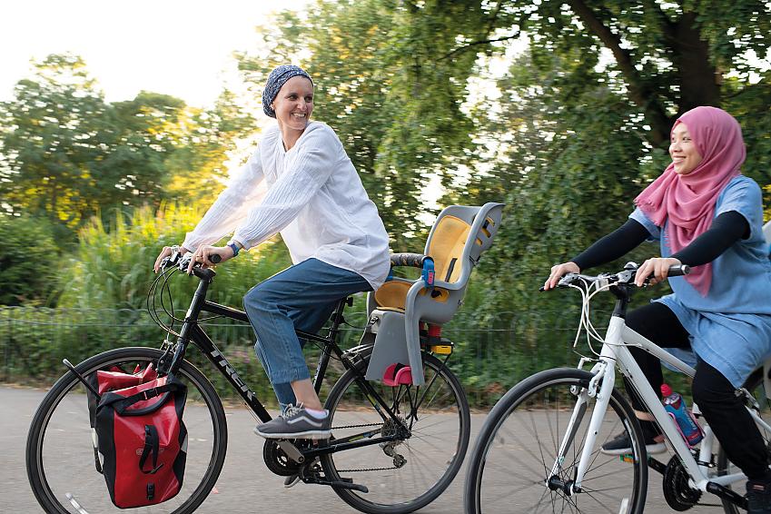 Two women on cycles. One is riding a black Trek with a child seat on the back. The other has  white bike. They're both wearing head scarfs and are smiling at each other