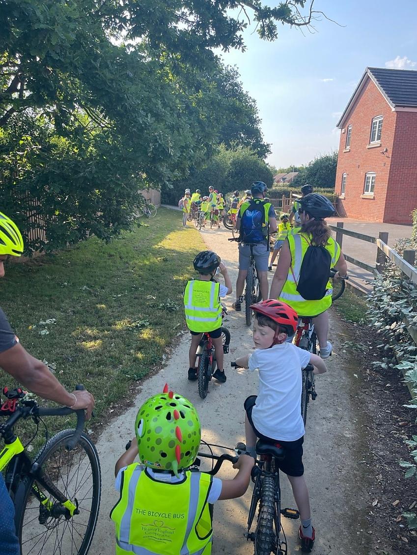 A line of kids and parents on bikes on a path getting ready to set off. They're wearing helmets and hi-vis jackets with 'The Bicycle Bus' on the back