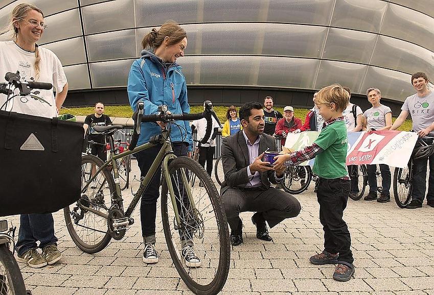 Humza Yousaf receiving a cake from a young boy with smiling cycle campaigners looking on