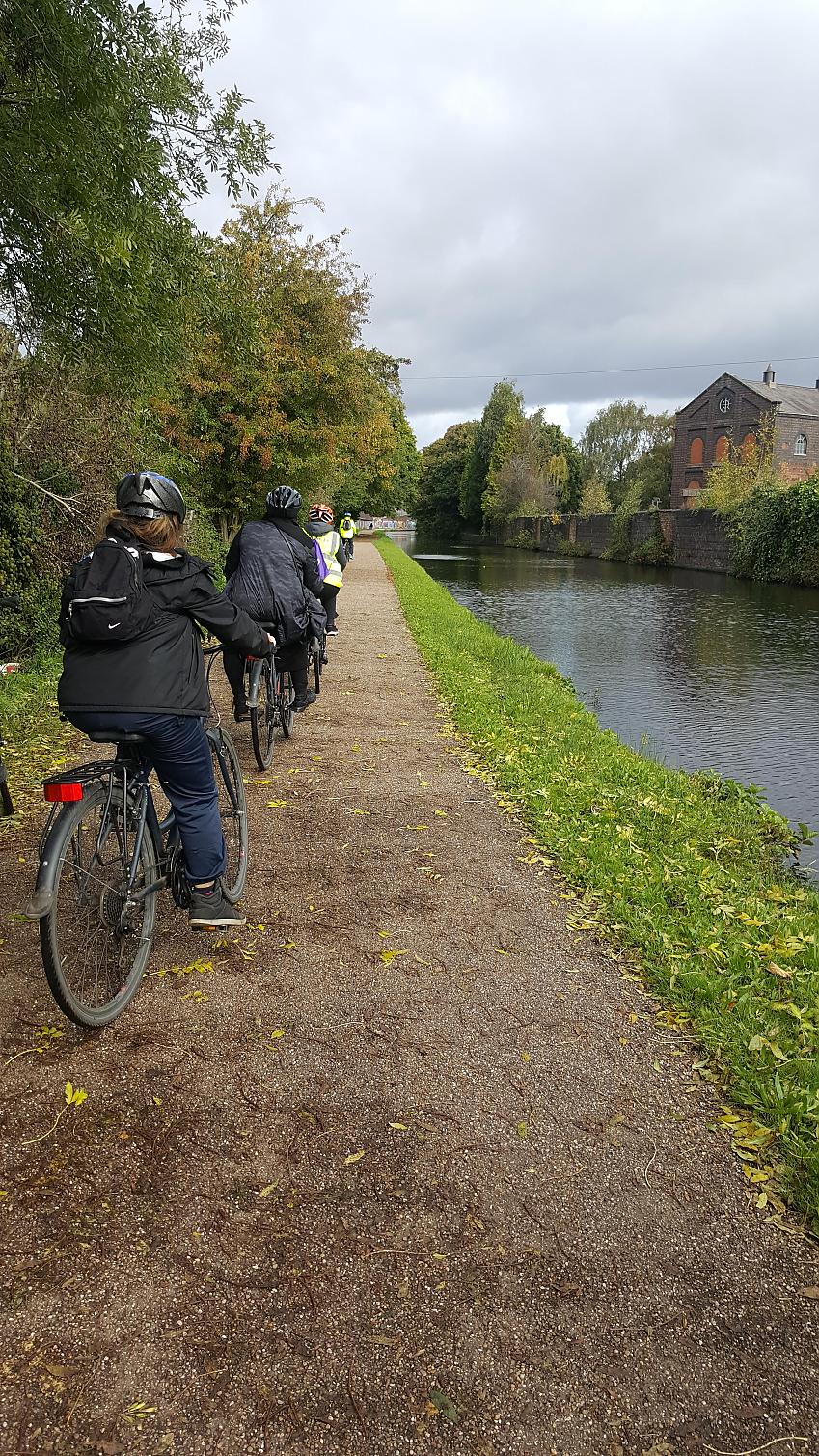 A group of people are cycling alongside a canal in single file. There are four of them. They wearing normal clothes, although two have hi-viz jackets. They all have helmets. It's autumn and the leaves are turning.