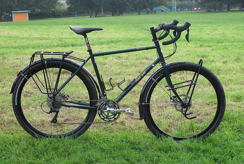 Spa Wayfarer, a dark green touring bike with drop handlebar, wide tyres, front and rear racks and mudguards. It's propped up in a park with trees and a playground in the background