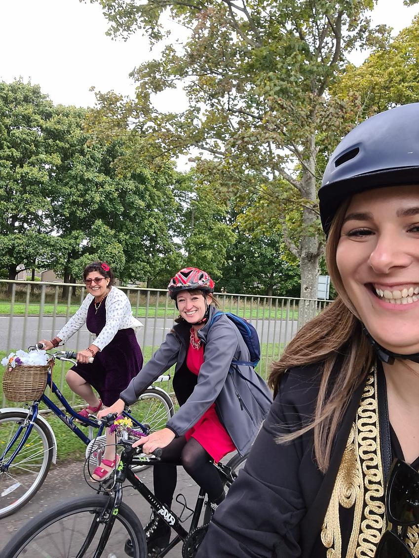 Three women are cycling and smiling at the camera. They are wearing smart clothing and have decorated their bikes with flowers