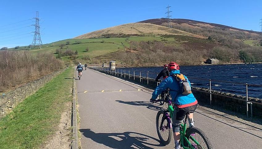 People cycle on a path beside a reservoir towards the hills