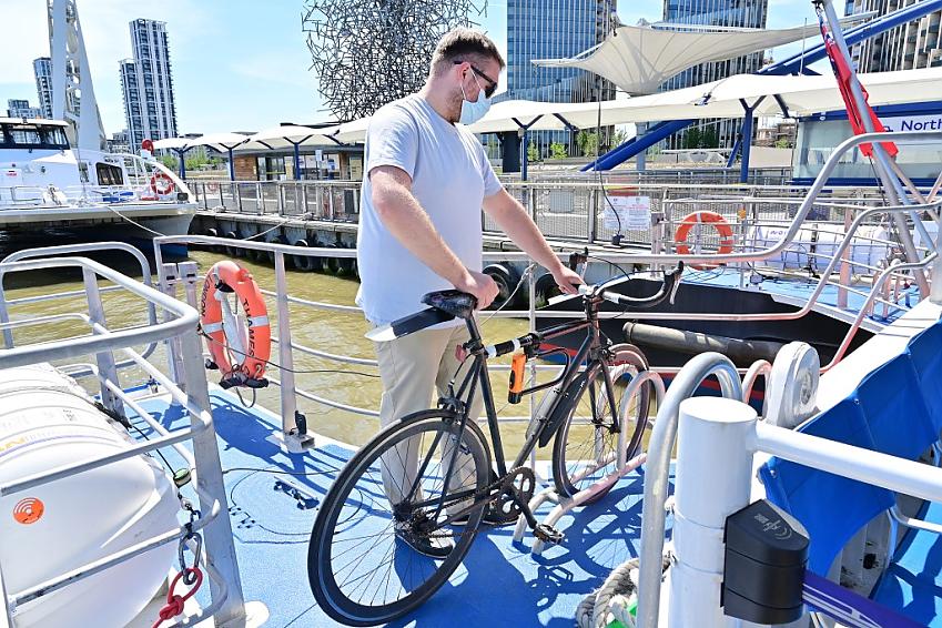 Person loads pedal bike onto bike rack on Thames Clippers boat on a sunny day.