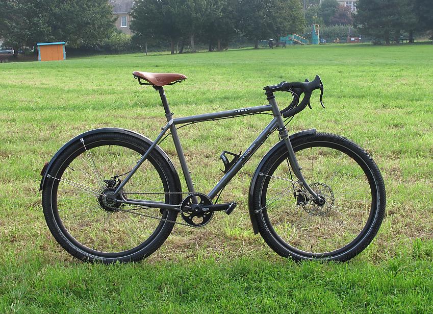 Thorn Nomad MK3, a dark steel grey touring bike with wide wheels, drop handlebar and mudguards. It's propped up in a park with trees and a playground in the background