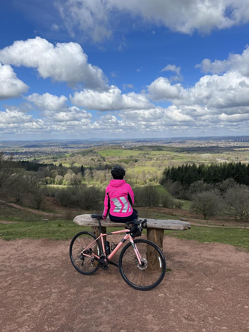 Alisha sits on a rustic wooden bench overlooking a countryside vista, her bicycle is leant on the bench behind her