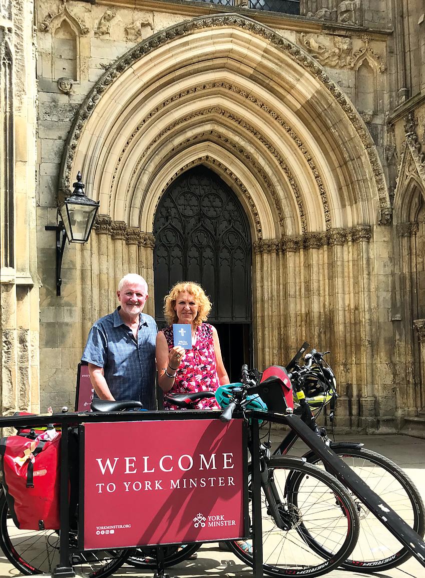 A man and woman stand with their bicycles in front of the large doors to a cathedral, a sign in front of them reads 'Welcome to York Minster'