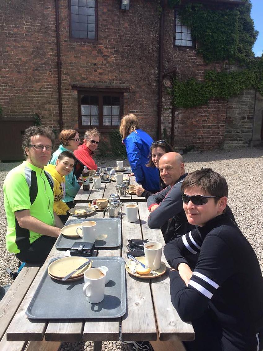 Cyclists are sitting at an outdoor table outside a National Trust property. They are tucking into cheese scones and mugs of tea