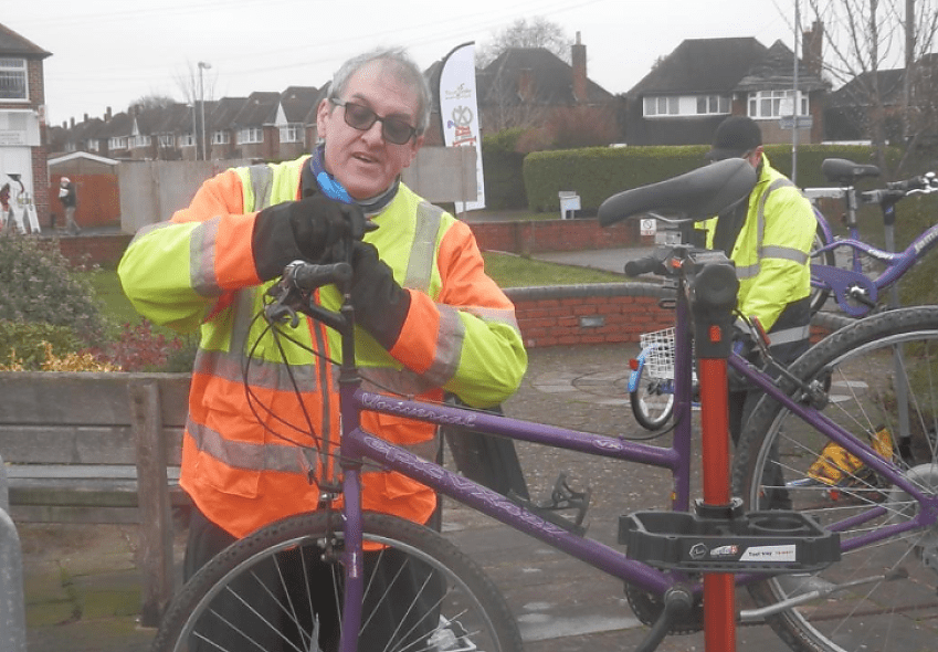 An older man in an orange and yellow jacket carries out repairs on a bicycle. Another person is stood behind him but not facing the camera