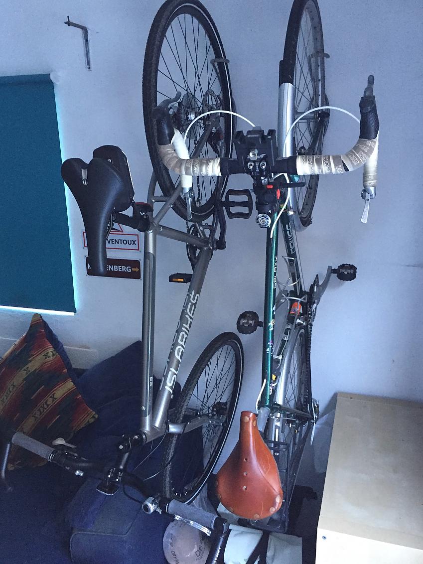 Two bikes are hung on a wall using brackets; they're hung nose to tail; one is a green road bike, the other silver hybrid
