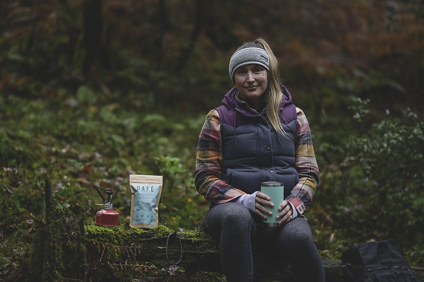 Rachael Walker sits on a mossy log. She is enjoying a mug of date coffee which she has just made using a camping stove. She is wearing warm clothing and smiling