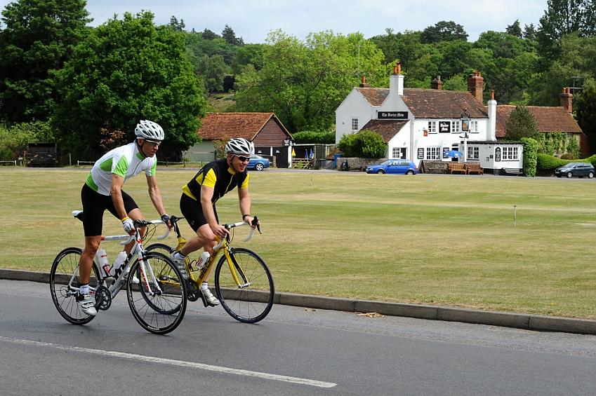 Two cyclists ride past a village green with the village pub in the background