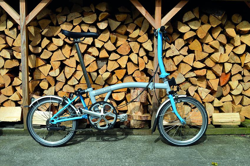 A light blue Brompton folding bike leaning against chopped wood in a wood shed, for some reason