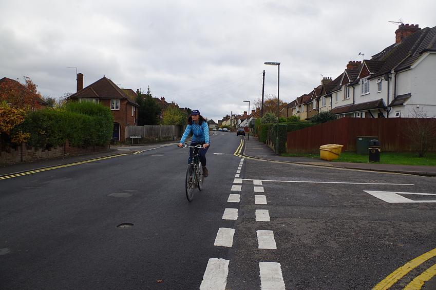 A woman is cycling past a junction very close to the white lines. She is wearing cap, blue jersey and blue leggings