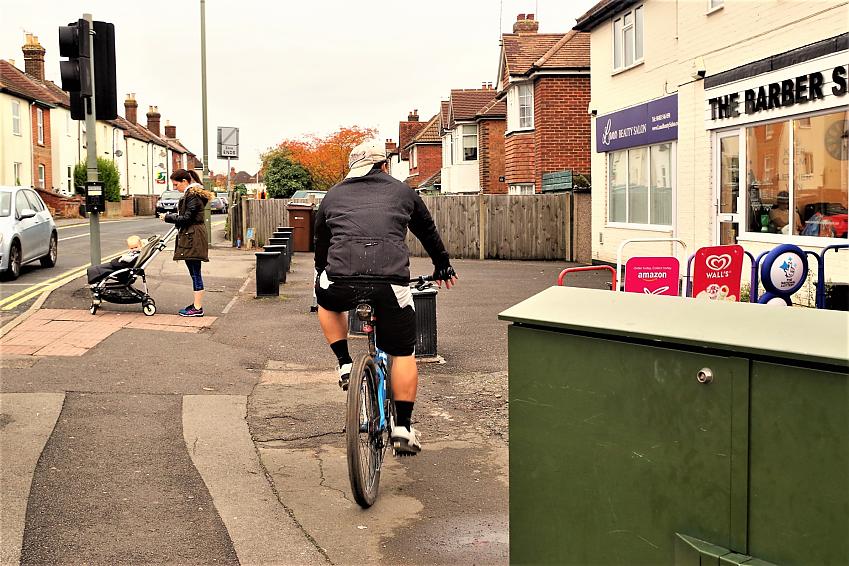 A cyclist is cycling along a path. There is a pedestrian with a buggy in front of him, along with bollards, bins and other hazards
