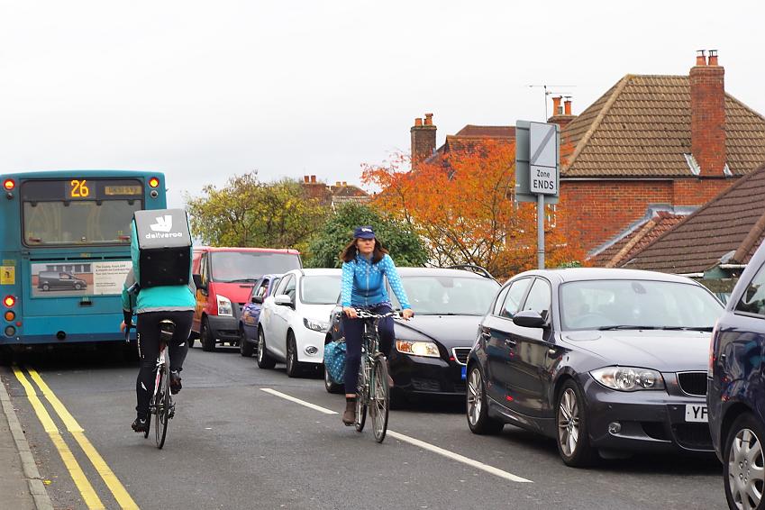 The same woman is riding along a busy road, next to a line of parked cars, with a deliveroo rider going past her in the opposite direction and a bus in the background