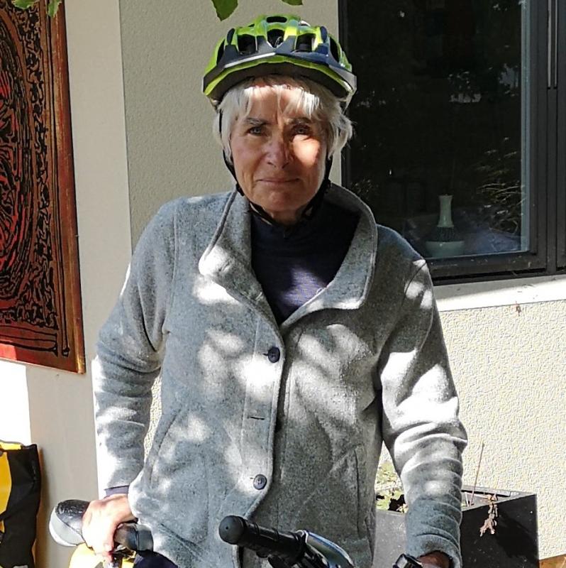 A woman is standing astride a grey hybrid bike with a packed trailer attached to it. She is wearing normal clothes: blue trousers and shoes and a grey fleece, as well as a cycle helmet.