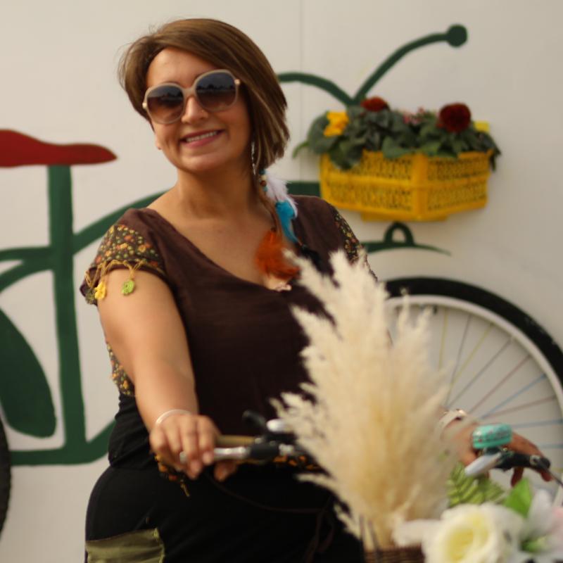 A woman is on a cream Dutch-style bike. The bike’s front basket is full of flowers. She is wearing leggings with a scarf tied around her waist and a brown T-shirt. She has on sunglasses and is smiling at the camera
