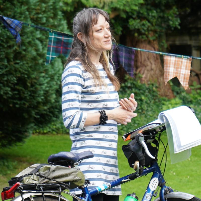 A woman is standing on a makeshift stage. She has a Dawes touring bike with her; an open notebook is balanced on the bike’s handlebars. She is wearing a blue and white striped top, black three-quarter-length trousers and cycling shoes. She is smiling.