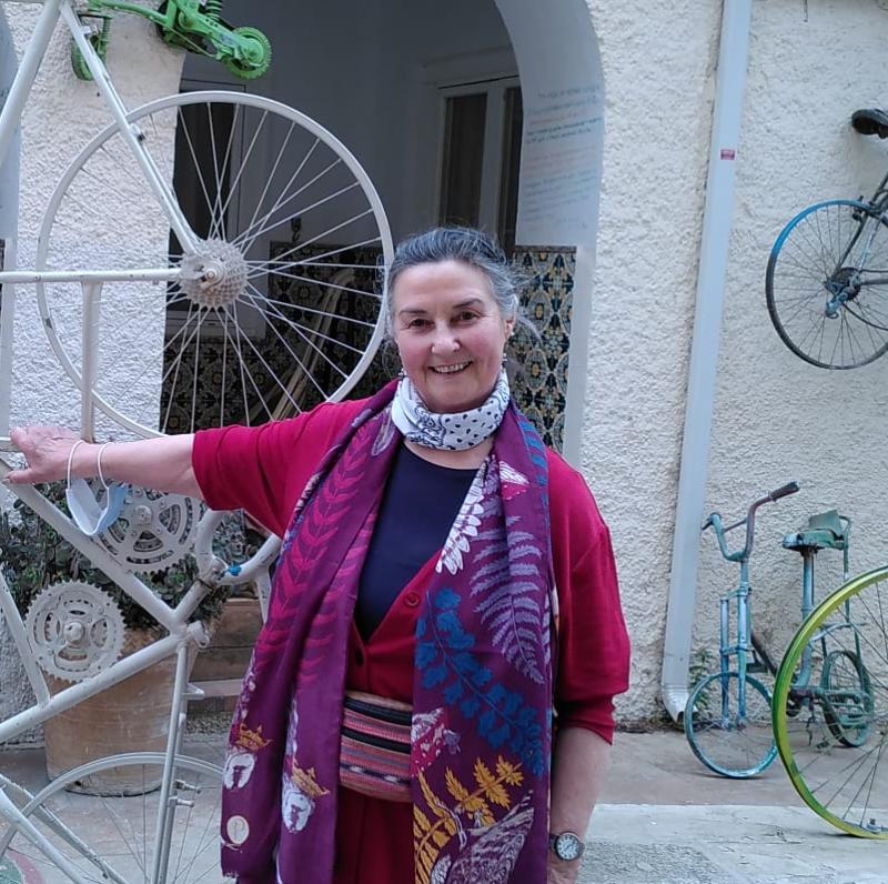 A woman is standing at the gates of a hotel in Spain. The building is covered with old bicycles and even the gates are made from bike frames. She is wearing black trousers, a bright cerise cardigan and blue top, as well as a brightly patterned scarf