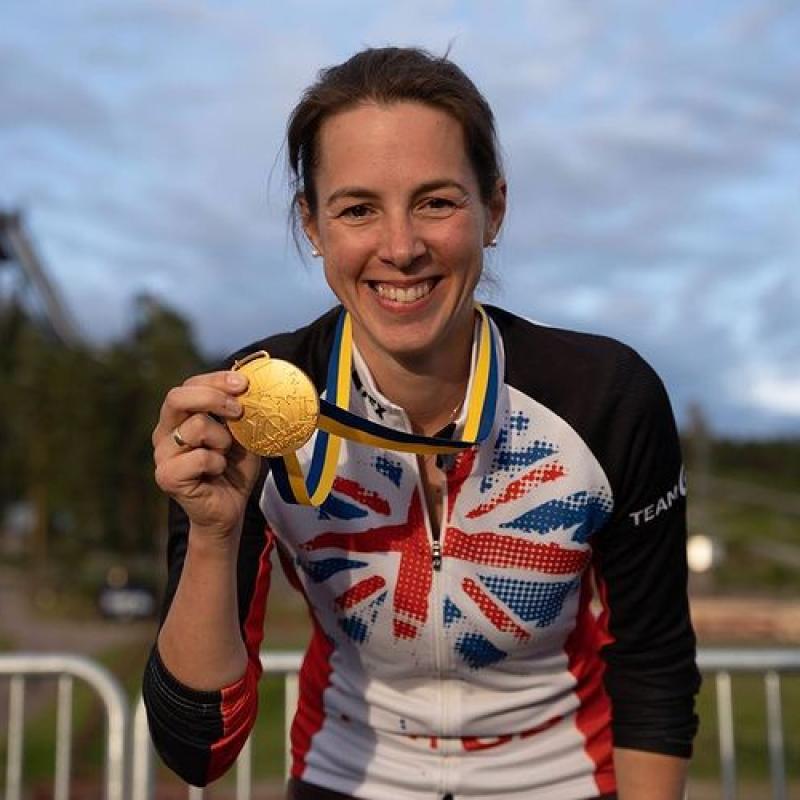 A woman in a GB cycling jersey is holding up a gold medal and smiling to the camera 